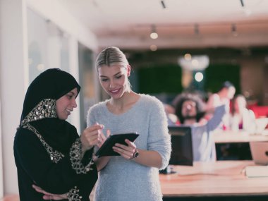 Bonde ve tesettürlü Müslüman iş kadını takım olarak çalışıyor ve tablet bilgisayarda birlikte çalışıyorlar. Arka planda çok ırklı meslektaşlar, modern birlikte çalışma ofisindeki farklı takımlar. 