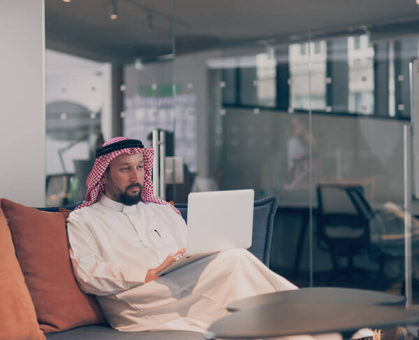 Mobile business communication and entrepreneurship. Arab businessman talking on smartphone and using a laptop computer, alone at open space coworking workplace or home office in the night. 