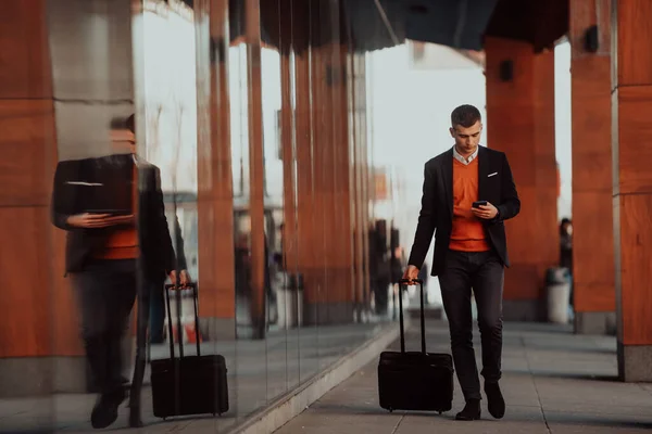 Going to airport terminal. Confident businessman traveler walking on city streets and pulling his suitcase drinking coffee and speaking on smartphone — стоковое фото