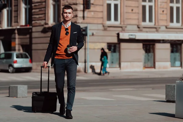 Voy a la terminal del aeropuerto. Viajero de negocios confiado caminando por las calles de la ciudad y tirando de su maleta bebiendo café y hablando en el teléfono inteligente — Foto de Stock