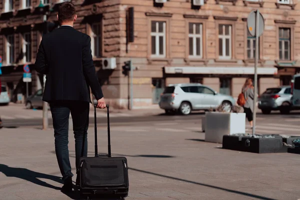 Jadę na lotnisko. Pewny siebie biznesmen podróżujący ulicami miasta, ciągnący walizkę, pijący kawę i mówiący na smartfonie — Zdjęcie stockowe