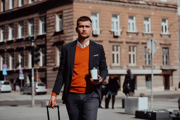 Voy a la terminal del aeropuerto. Viajero de negocios confiado caminando por las calles de la ciudad y tirando de su maleta bebiendo café y hablando en el teléfono inteligente — Foto de Stock