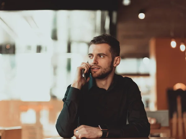 Mladý podnikatel v černé košili sedí v kavárně a používá smartphone během přestávky na kávu. — Stock fotografie