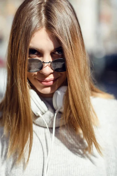 A modern girl with sunglasses walking down the street listens to music in headphones. — стоковое фото