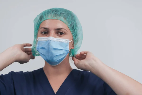 여성 의사나 과학자를 가까이 서 마스크를 쓰고 회색 배경에 수술 모자를 씌운 채로 요. 마스크를 조정하고 있어요 — 스톡 사진