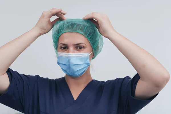 Close up of female doctor or scientist with a medical mask and surgical cap over grey background. She is adjusting mask with — Stock Photo, Image