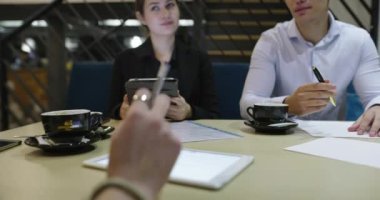 Bir grup iş arkadaşı kafeteryada buluşuyor, teknolojiyle çalışıyor ve yakın planda kahve içiyor.