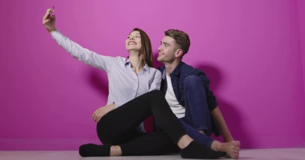 ピンクを背景にスマートフォンで自撮り写真を撮りながら 楽しいカラフルな服を着た正のカップルの肖像 — ストック動画