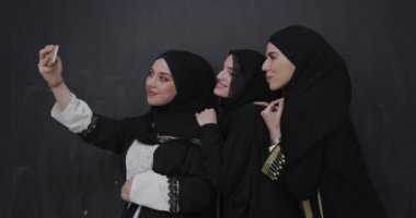 Müslüman kadınlar tesettürlü şık bir elbise giyip tesettürlü arka plan üzerinde akıllı telefon kullanarak selfie çekiyorlar.