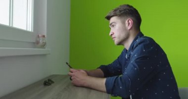 Gülümseyen yakışıklı genç adam evdeki yeşil duvarın üzerinde akıllı telefon kullanıyor.