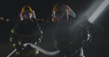 İtfaiyeci erkek ve kadın grup acil bir durumda alevlerle savaşmak için su kullanıyor. İtfaiyeci ekibi yangın geçirmez ekipmanla gece eğitiminde itfaiye aracının yanında. Yavaş çekim. 