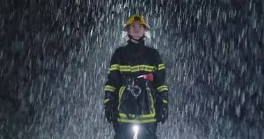 Kahraman fotoğrafı, yağmurun üstünde koruyucu kaskını takmış ve itfaiyeci üniformasıyla kameraya bakan yorgun Amerikalı kadın itfaiyecinin portresi. Yüksek kalite 4K görüntü. Yavaş çekim.