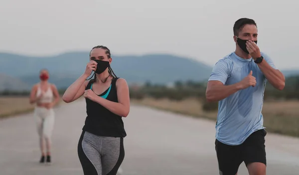 多民族跑步者团体戴着口罩跑步，与外界保持距离。为了安全起见，健康的多样化队伍在晚上穿着运动服在大自然的跑道上慢跑. — 图库照片