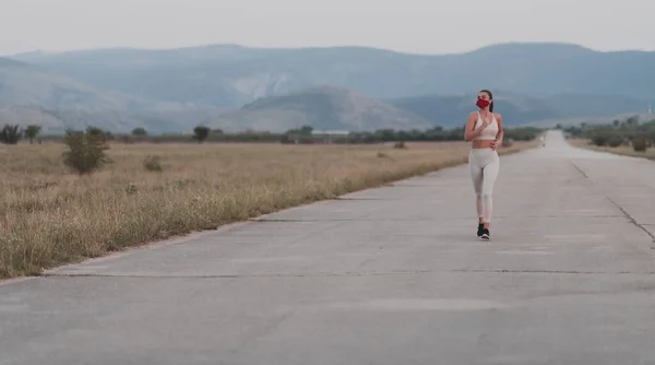 Determined fitness woman in short clothes wearing red protective face mask running outdoors in the city during coronavirus outbreak. Covid 19 and physical jogging activity sport and fitness.