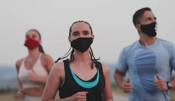 Wieloetniczni biegacze noszą maski na twarzy. Utrzymują dystans społeczny na zewnątrz. Odpowiedni zdrowy zróżnicowany zespół nosi odzież sportową jogging w godzinach wieczornych na torze sportowym przyrody odległości dla bezpieczeństwa. — Zdjęcie stockowe