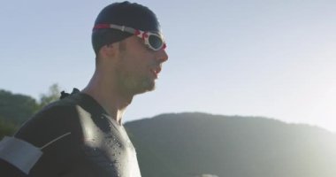 Kararlı triatlon atletinin yavaş çekim portresi hazırlanıyor ve güneş doğarken gölde yüzme eğitimi için gözlük şapkası ve dalış kıyafeti hazırlıyor. Seçici odak. 