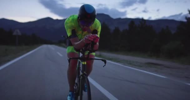 プロのレーシングバイクに乗ってトライアスロン選手の夜の自転車トレーニング 夕方には曲がりくねった田舎道で健康的な強烈なワークアウト スローモーショントラッキングショット — ストック動画