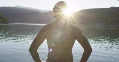 Dalış kıyafetli profesyonel triatlon sporcusu Demir Adam yarışması için yüzme antrenmanına bir gölün su sıçratan sularında koşup zıplayarak başlıyor..
