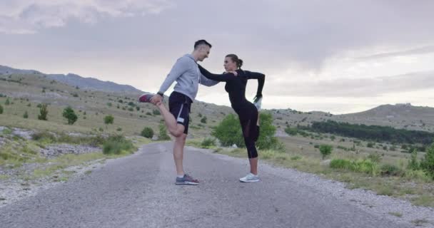 Couple of determined athletes stretching together, warming up and preparing for running training on a panoramic road. — Stockvideo