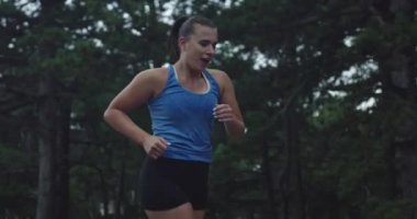 Ağır çekimde, spor giysili genç bir kadının, yağmur altındaki bir kır yolunda, yemyeşil bir ormanda koşarkenki görüntüsü..
