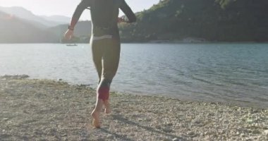Dalgıç kıyafetli profesyonel triatlon sporcusu demir adam yarışması için yüzme antrenmanına bir gölün su sıçratan sularında koşup zıplayarak başlıyor. Yavaş çekim izleme çekimi. 