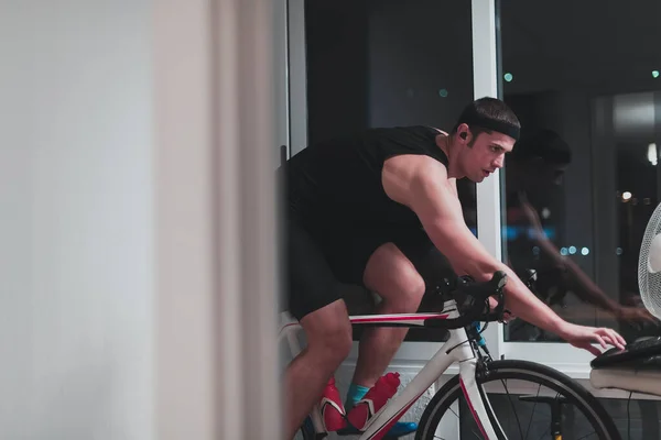 Man cycling on the machine trainer he is exercising in the home at night playing online bike racing game — Stock Photo, Image
