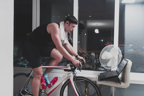 Hombre ciclismo en el entrenador de la máquina que está ejercitando en el hogar por la noche jugando juego de carreras de bicicletas en línea —  Fotos de Stock