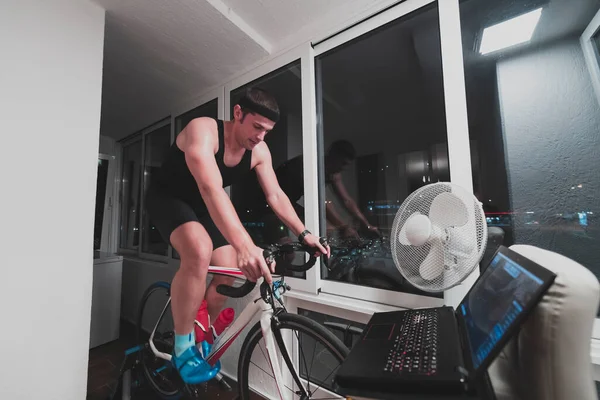 Man cycling on the machine trainer he is exercising in the home at night playing online bike racing game — Stock Photo, Image