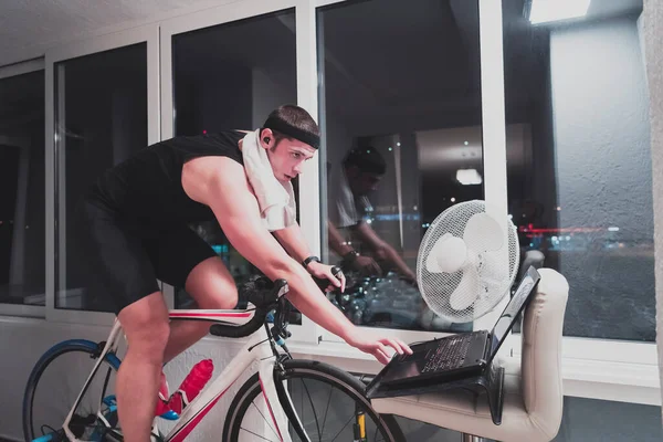 Hombre ciclismo en el entrenador de la máquina que está ejercitando en el hogar por la noche jugando juego de carreras de bicicletas en línea —  Fotos de Stock