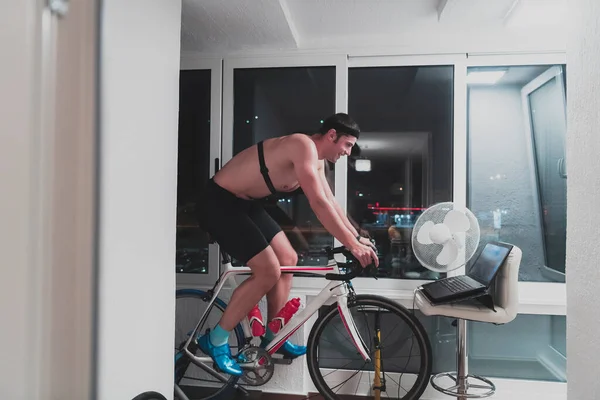 Homme vélo sur l'entraîneur de la machine, il fait de l'exercice à la maison la nuit en jouant en ligne jeu de course de vélo — Photo