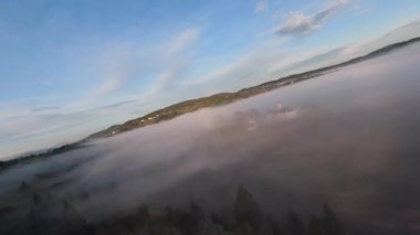 Sabah sisli orman üzerinde uçarken, gün doğumunda ağaçların tepesinde sis ve güneş ışınları vardı. FPV insansız hava aracının, dağlık tepe arazili doğa manzarası üzerindeki görüntüsü. Sinematik görünüm.