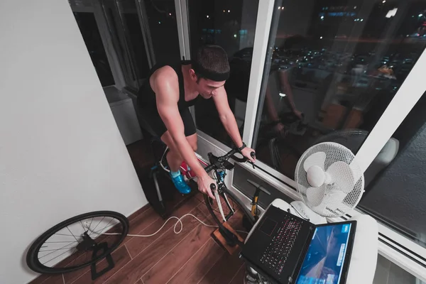 Hombre ciclismo en el entrenador de la máquina que está ejercitando en el hogar por la noche jugando juego de carreras de bicicletas en línea —  Fotos de Stock