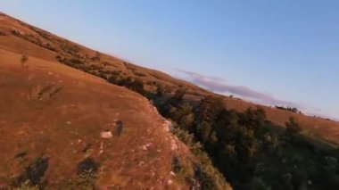 Gün batımında, dağ tepelerinin üzerinden aşırı yakın uçuş. Fpv spor insansız hava aracının dağlık tepe arazili güneşli manzarası. Dinamik sinematik görünüm.