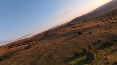 Gün batımında, dağ tepelerinin üzerinden aşırı yakın uçuş. Fpv spor insansız hava aracının dağlık tepe arazili güneşli manzarası. Dinamik sinematik görünüm.