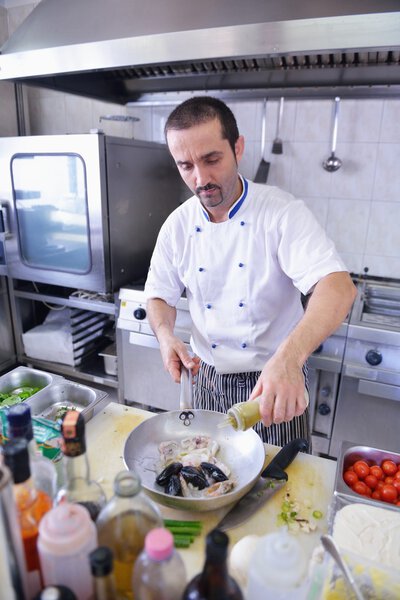 Handsome chef dressed in white uniform making pasta salad and seafood in modern kitchen