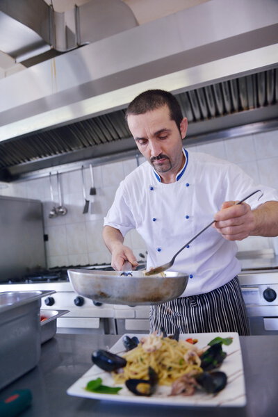 Handsome chef dressed in white uniform decorating seafood pasta salad in modern kitchen