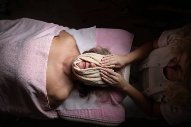 Modern bir kaplıcadaki profesyonel bir kadın terapistten rahatlatıcı bir yüz masajı alan olgun bir kadına yakın çekim. Müşteri bir tedavi yatağında havlu ve saç bandıyla yatıyor, cilt bakımının keyfini çıkarıyor.