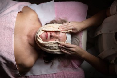 Modern bir kaplıcadaki profesyonel bir kadın terapistten rahatlatıcı bir yüz masajı alan olgun bir kadına yakın çekim. Müşteri bir tedavi yatağında havlu ve saç bandıyla yatıyor, cilt bakımının keyfini çıkarıyor.