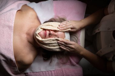 Modern bir kaplıcadaki profesyonel bir kadın terapistten rahatlatıcı bir yüz masajı alan olgun bir kadına yakın çekim. Müşteri bir tedavi yatağında havlu ve saç bandıyla yatıyor, cilt bakımının keyfini çıkarıyor.