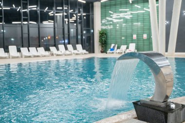 Temiz mavi suyu olan çağdaş bir yüzme havuzu şık bir ışıklandırma ve beyaz şelale sandalyeleriyle çevrili metal bir şelale. Refah, lüks, rahatlama ve modern kaplıca kavramı