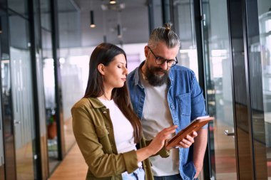 İki iş adamı, parlak bir modern ofis koridorunda bir tablet üzerindeki bilgileri incelerken, takım çalışması, iletişim ve dijital işbirliğini sembolize ediyorlar.