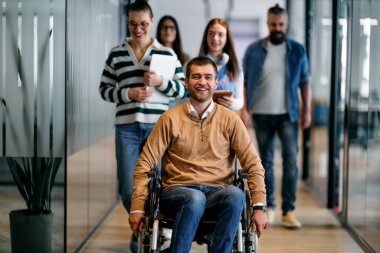 Bir grup iş adamı modern ofis koridorunda birlikte yürüyor. Tekerlekli sandalyedeki güler yüzlü adam tarafından yönetiliyor. Kapsama, eşitlik ve takım çalışmasını temsil ediyor.
