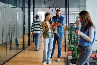 İki iş adamı, parlak bir modern ofis koridorunda bir tablet üzerindeki bilgileri incelerken, takım çalışması, iletişim ve dijital işbirliğini sembolize ediyorlar.