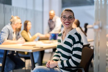 Gülümseyen iş kadını modern bir ofiste güvenle otururken, ekibi arka planda işbirliği yaparak, olumlu, takım çalışması, liderlik ve kurumsal bir ortamda kariyer başarısını temsil ediyor.