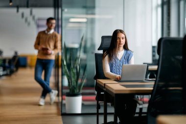 Meslektaşım arka planda, profesyonel bir ortamda üretkenliği, odaklanmayı ve takım çalışmasını temsil ederken, masa başında oturan ve parlak, modern bir ofiste çalışan bir iş kadını.