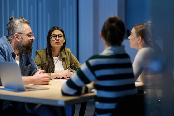 Группа разнообразных коллег, которые ведут серьезную дискуссию за столом переговоров, во главе с бородатым менеджером-мужчиной, представляя командную работу, лидерство и стратегию развития современного бизнеса