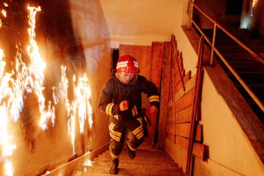 Bir itfaiyeci acil bir durum sırasında yanan merdivenlerden aşağı koşuyor. Koruyucu giysi, kask ve üniforma giyiyor. Tehlikeli bir operasyonda yangın güvenliği ve kurtarma operasyonu gösteriyor.