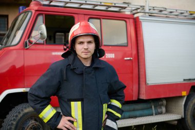 Tam teçhizatlı genç bir itfaiyecinin portresi. Kırmızı kasklı ve koruyucu üniformalı. İtfaiye aracının önünde duruyor. Bu resim acil servisleri göstermek için idealdir