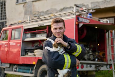 Bir itfaiyeci kurtarılmış bir kediyi itfaiye arabasının önünde tutuyor. Bu görüntü cesaret, şefkat ve itfaiyecilerin hem insan hem de hayvan hayatları kurtarmada oynadıkları önemli rolü temsil ediyor. Mükemmel.