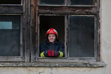 Bir itfaiyeci gülümser ve yıpranmış eski bir pencereden dışarı bakar. Kırmızı bir kask ve koruyucu eldiven takıyor. Şehir ortamında kurtarma ve güvenliği temsil ediyor..